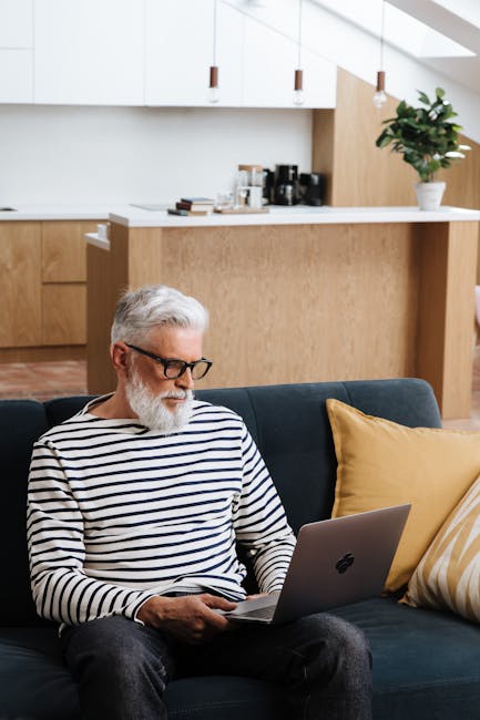 Elderly man with eyeglasses using a laptop in a cozy, modern living room.